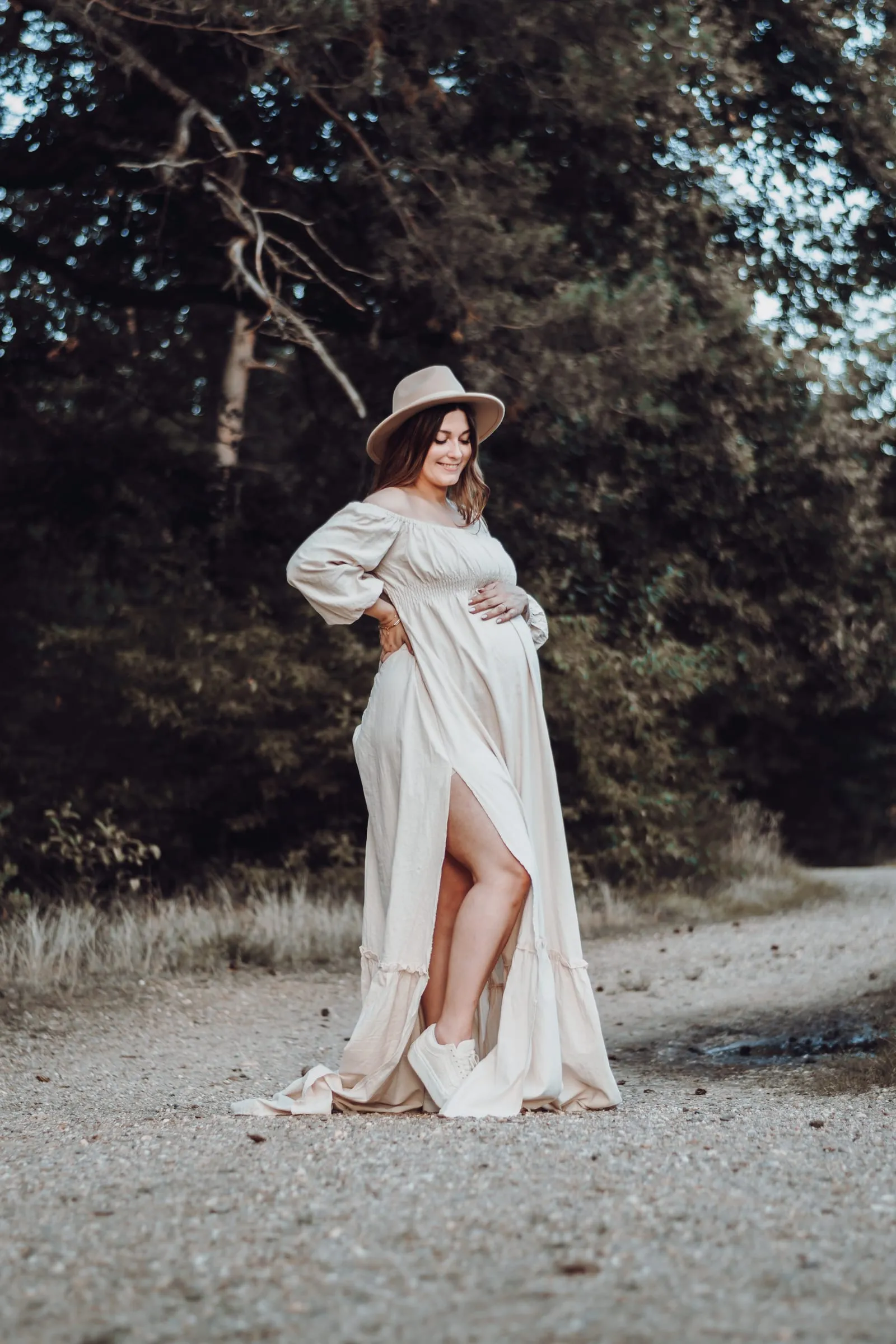 Schwangere im weißen Kleid mit Hut auf einem Waldweg