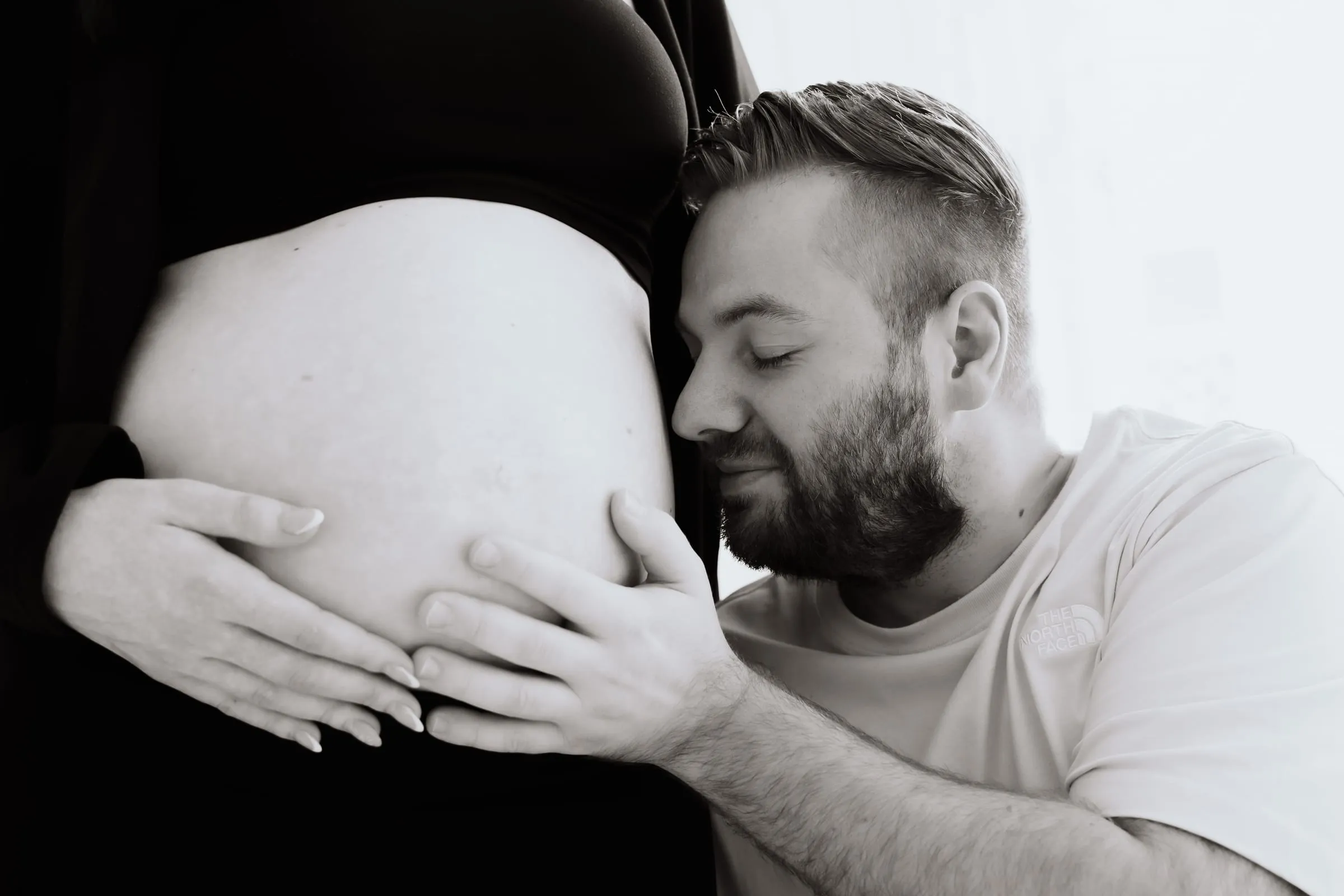Der werdende Vater schmiegt sein Gesicht an den nackten Babybauch, ihre Hände liegen auf seinen, in Schwarzweiß