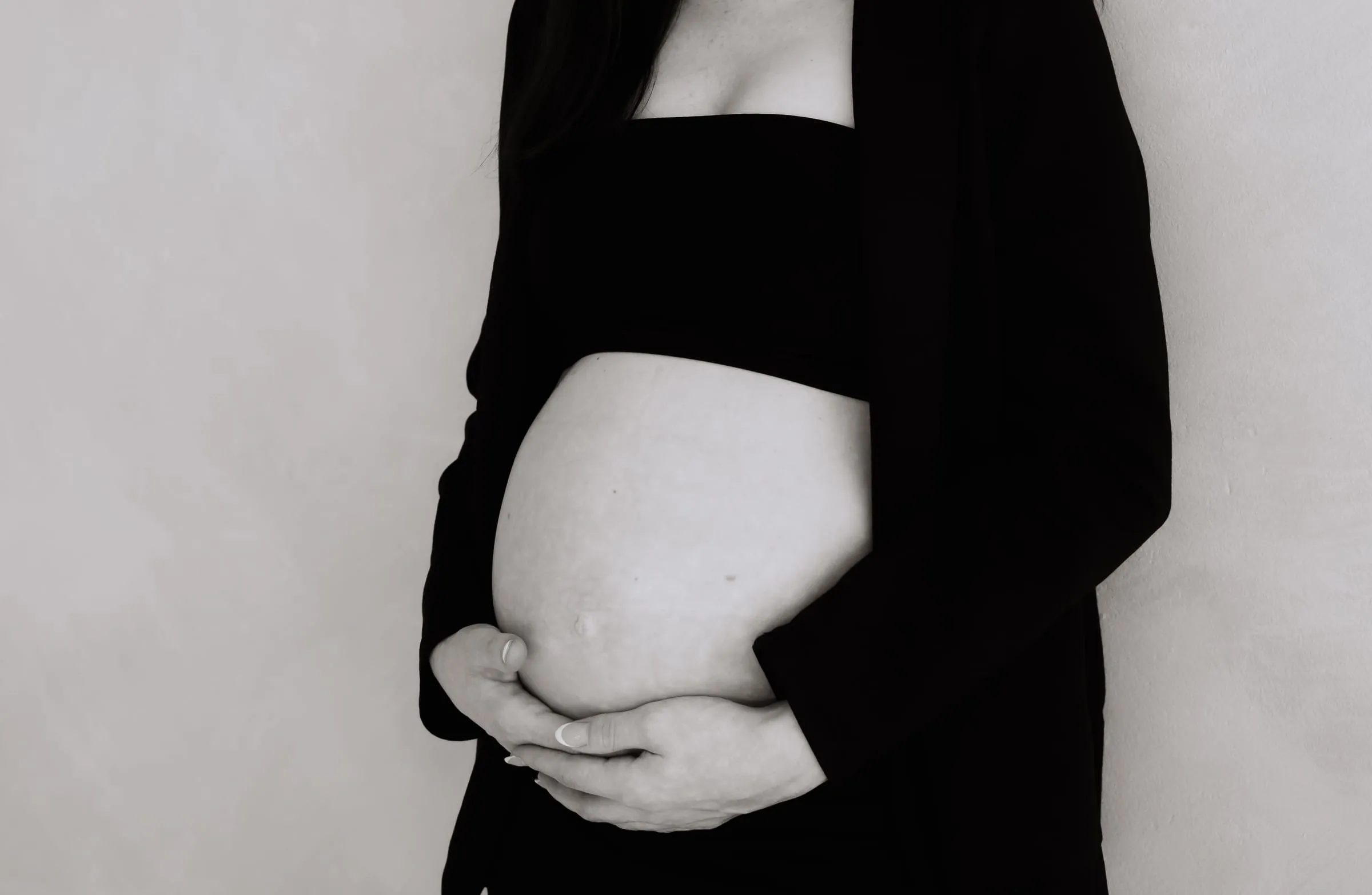 Nahaufnahme des Babybauchs zwischen den offenen Seiten eines schwarzen Blazers, beide Hände darunter