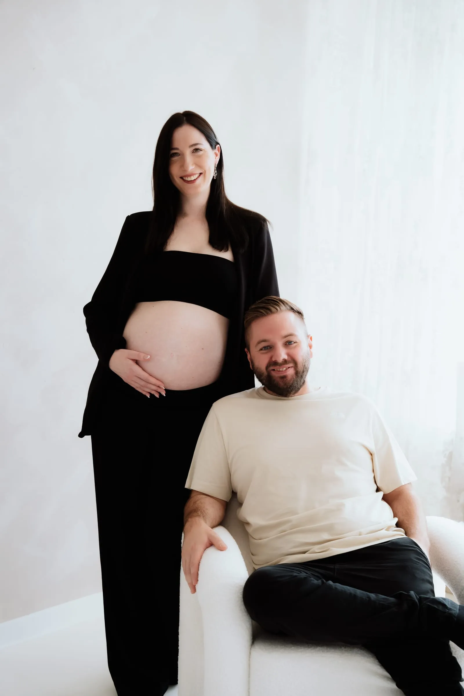 Die Schwangere steht neben ihrem Partner, der in einem weißen Sessel sitzt, beide lachen in die Kamera