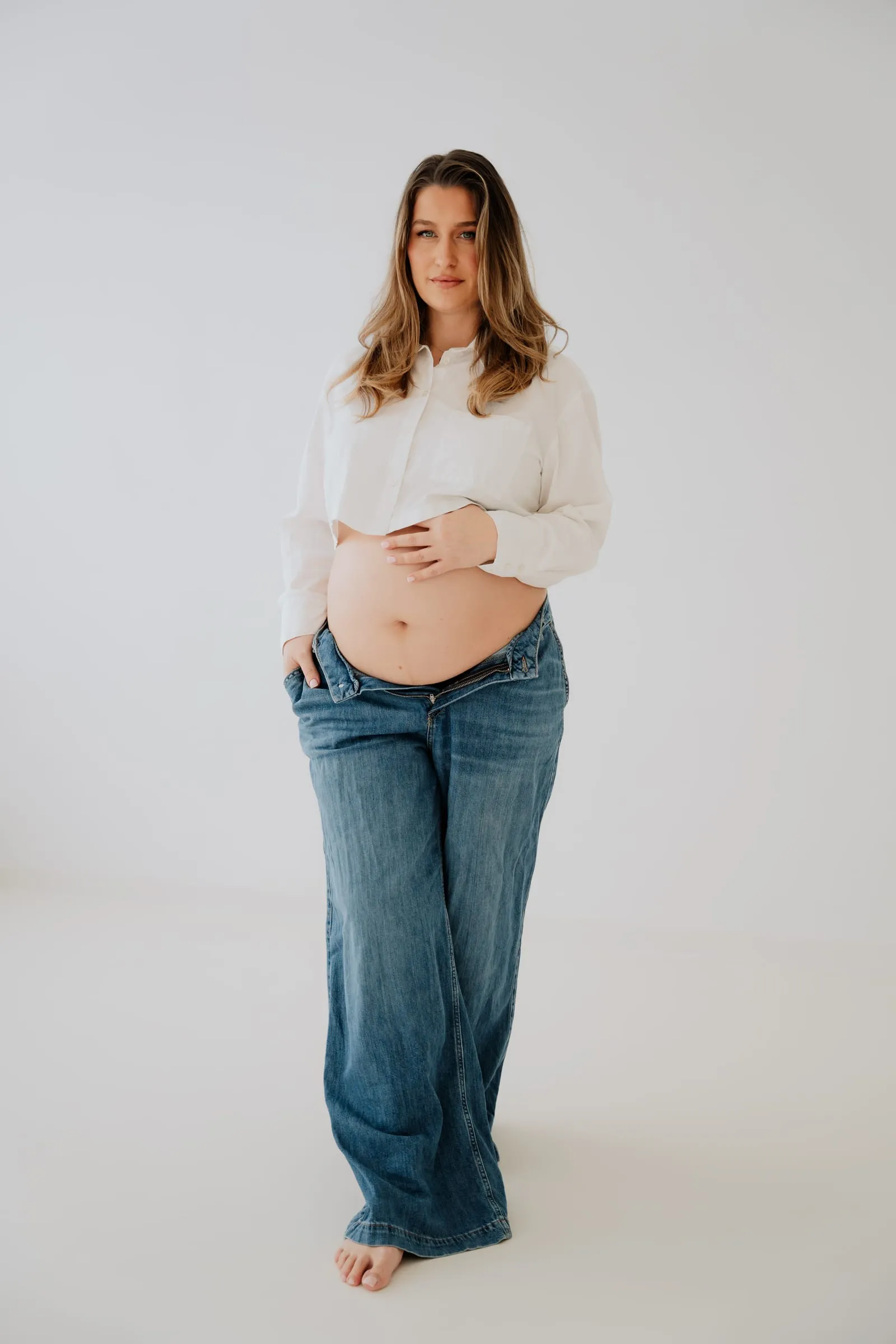 Schwangere in weißer Bluse und weiter Jeans steht barfuß im hellen Studio, eine Hand am Babybauch