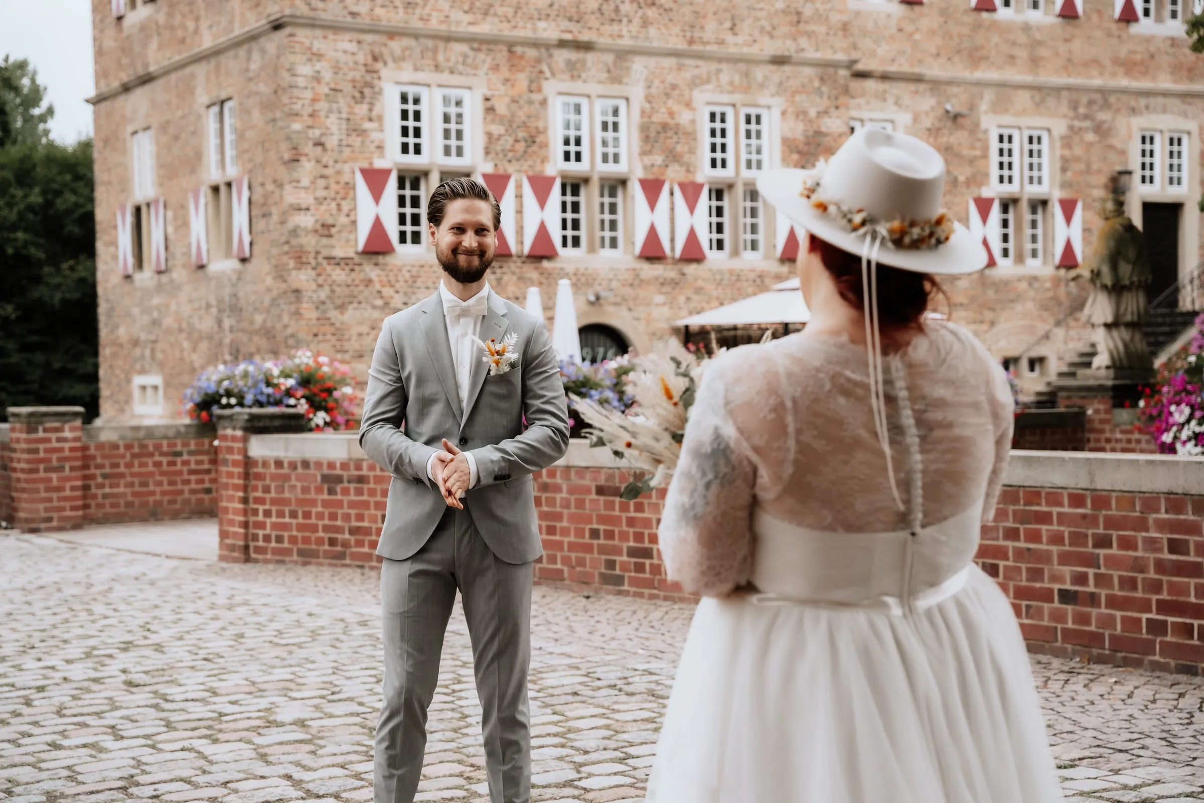 Das Brautpaar lacht sich im Schlosshof an, sie mit Hut und Tüllrock