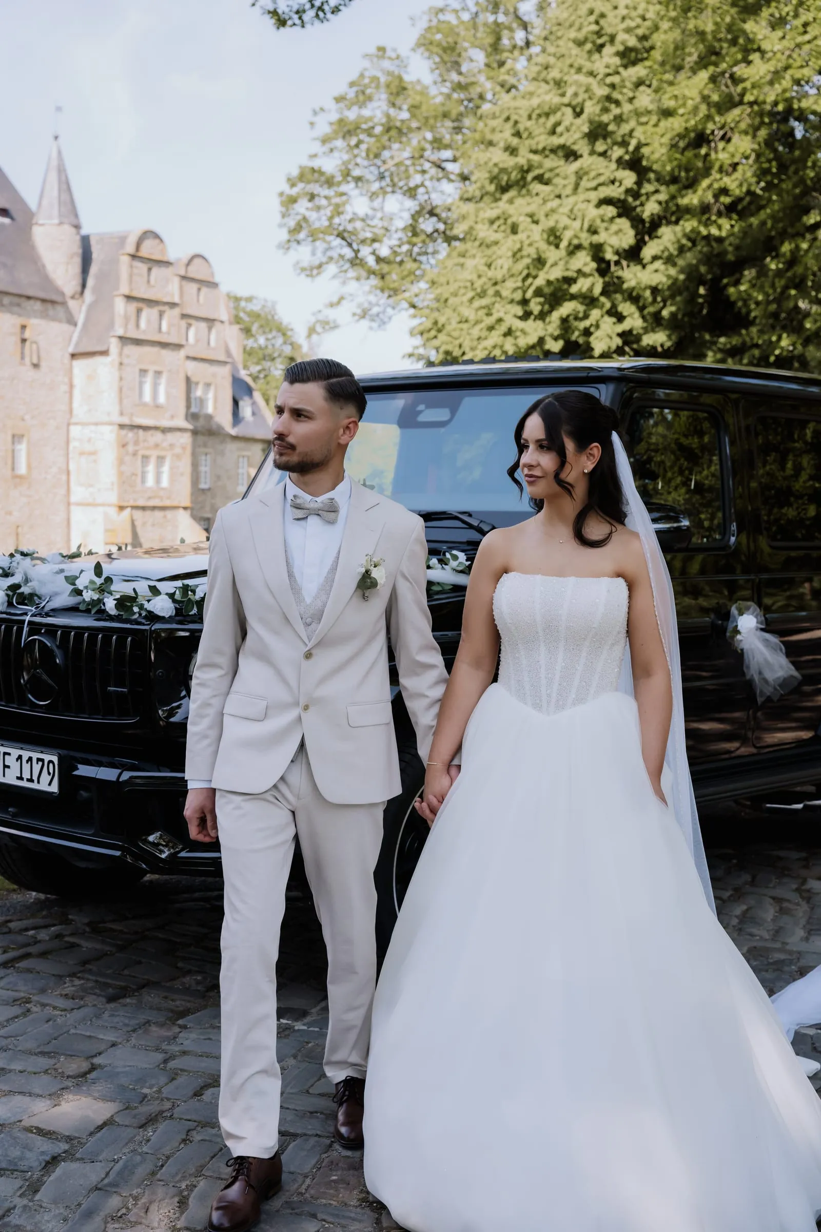 Das Brautpaar Hand in Hand vor dem Schloss, daneben der Wagen