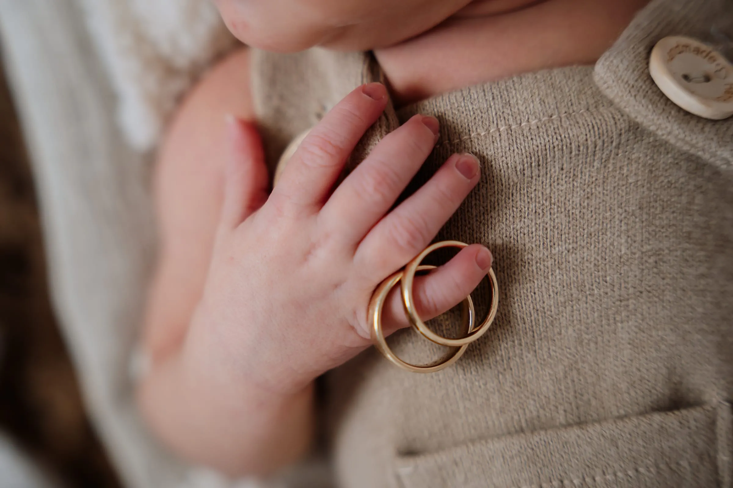 Die Hand des Babys auf beigem Strick, daneben die Eheringe der Eltern