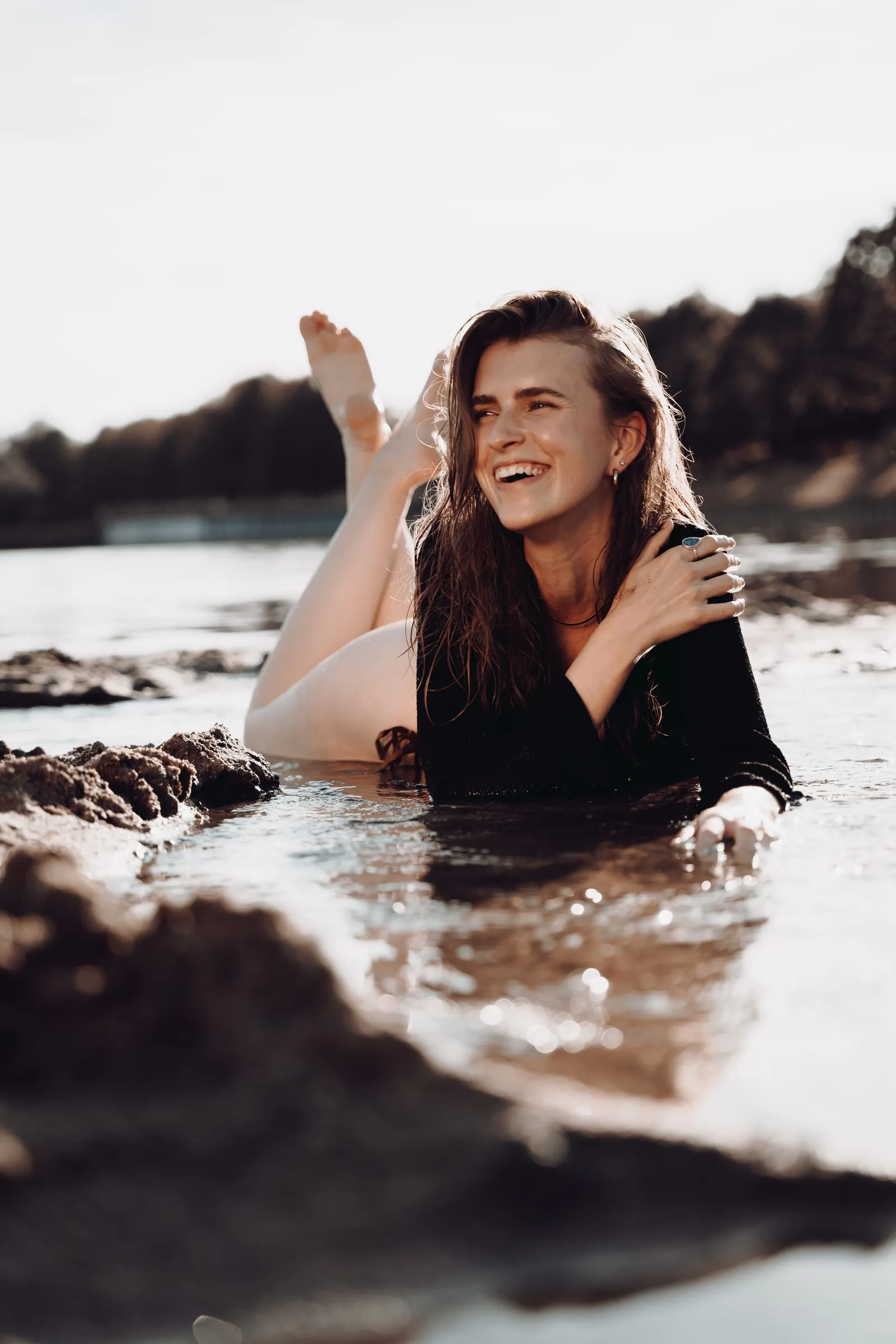 Junge Frau liegt lachend im flachen Wasser am Ufer, die Füße in der Luft, im Gegenlicht