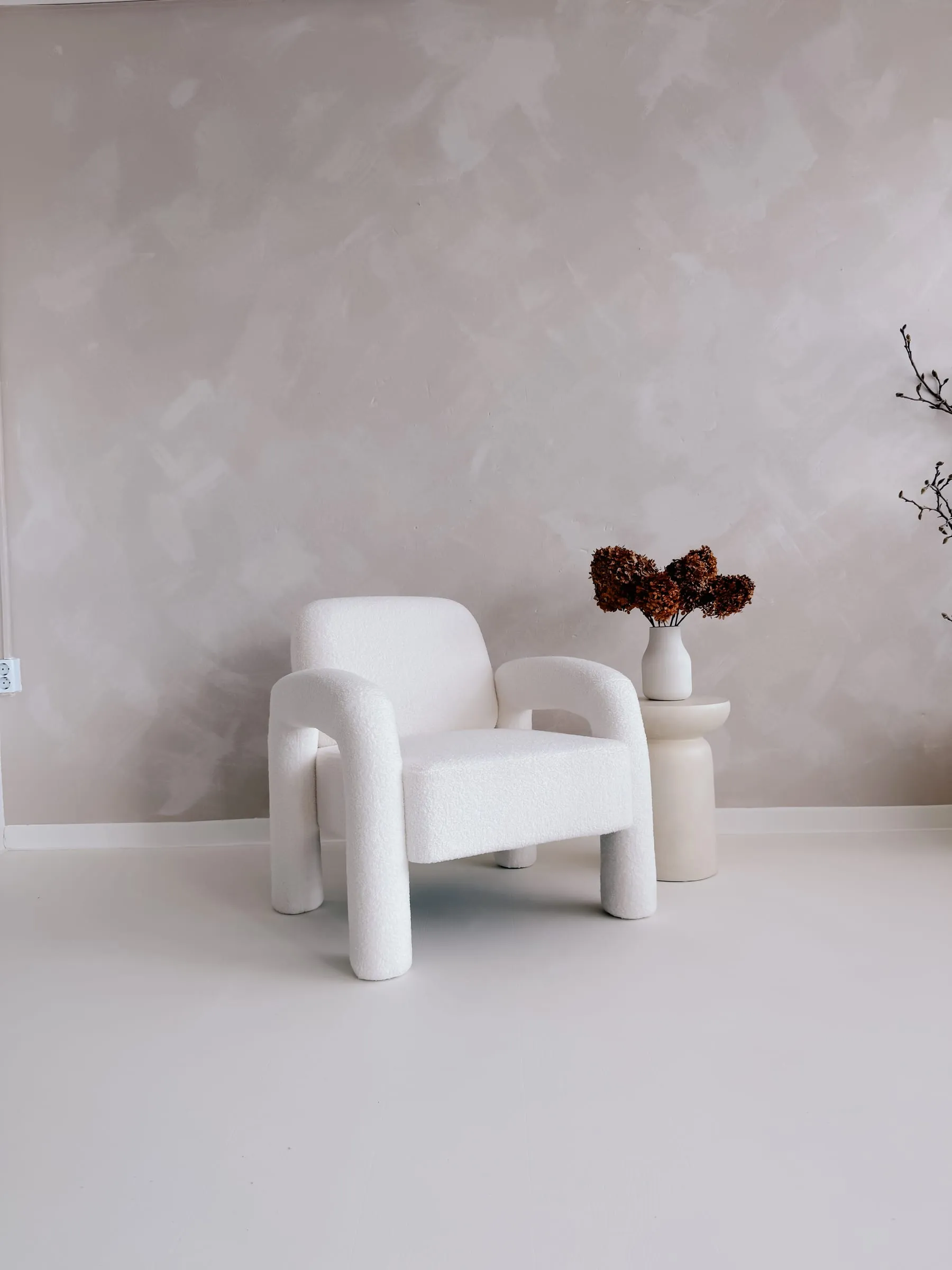 Weißer Bouclé-Sessel vor einer hellen Putzwand, daneben eine Vase mit getrockneten Hortensien